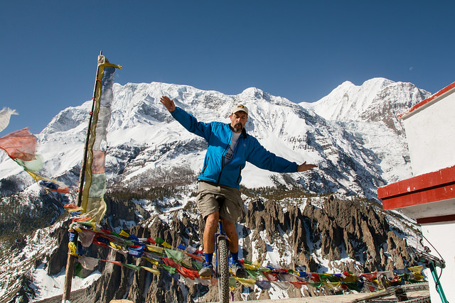 A man balances on a unicycle with his arms extended in a mountain landscape. The mountain behind him is snow-capped. Himalayan prayer flags flap in the breeze.