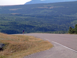 A lone woman rides a bike up a hill in a vast landscape. Behind her is a valley with scrub and trees; a mountain looms behind that. The air looks hazy.