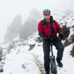 A mountain unicyclist wearing cold-weather gear rides down a snow path in the mountains. The peaks behind him are partially occluded by clouds. The landscape is substantially rocky. He is smiling broadly.