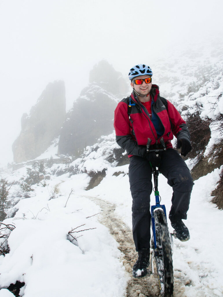 A mountain unicyclist wearing cold-weather gear rides down a snow path in the mountains. The peaks behind him are partially occluded by clouds. The landscape is substantially rocky. He is smiling broadly.