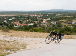 A bicycle with one pannier is parked on the side of a road, on a hillside overlooking a valley. The valley is dotted with farmhouses with red tile roofs.