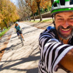A smiling cyclist wearing a black and white striped shirt and a helmet takes a selfie on a path lined with autumnal trees. Out of focus behind him is seen a smiling woman on a hybrid bike.