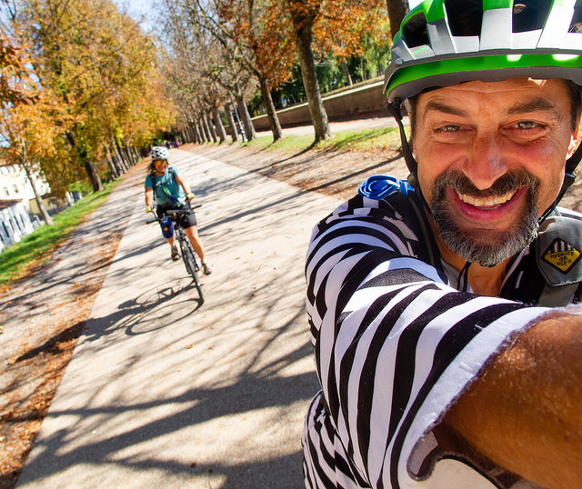 A smiling cyclist wearing a black and white striped shirt and a helmet takes a selfie on a path lined with autumnal trees. Out of focus behind him is seen a smiling woman on a hybrid bike.