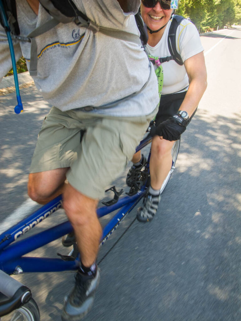 A pair of cyclists ride a blue Cannondale tandem bike. The captain appears to be holding the camera in his outstretched arm; the image shows his muscular legs, appearing to be moving quickly because the image is motion blurred. The stoker, a woman wearing Keen sandals, is smiling at the camera. 