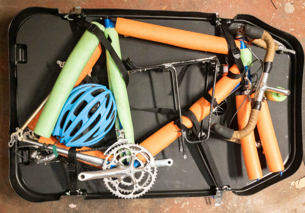 A top-down view of an open hard-shell bicycle travel case packed with a road bike. The handlebars are detached, a cargo rack and helmet are nestled in the middle of the frame, and the frame tubes and fork are protected with green and orange pool noodles. The bike is strapped to the sides of the case.