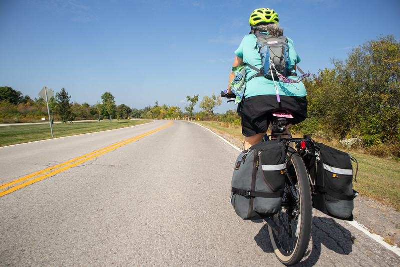 A woman on a touring bicycle rides on a flat, open road with a grassy verge. Trees in the distance show some fall colors. The photo is shot from the level of the rear tire, giving an impression of movement. The woman has ribbons and a colorful bandana attached to her backpack, blowing in the breeze. The weather is sunny, with a few wispy clouds visible.