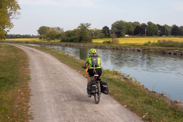 A woman on a bicycle with rear panniers rides on a gravel pathway along a waterway on a pleasant-looking day. Across the waterway is a field of yellowing plants, possibly soybeans.