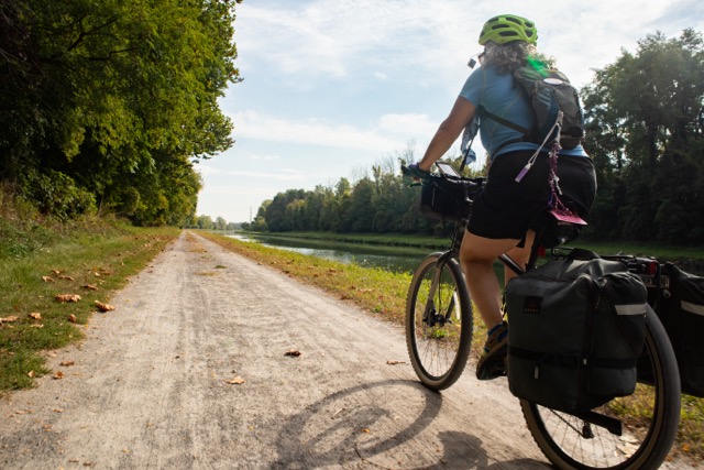 A woman on a bicycle with panniers rides on a gravel pathway by a canal. The weather is partly sunny; both sides of the canal are lined with trees. It looks pleasant.