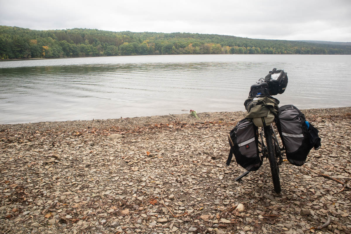 A gravel bicycle with a front handlebar bag, two large rear panniers, and a tent strapped to the rear rack, is parked on a rocky beach on a lake. The weather is mostly gray, with some small breaks in the clouds. Across the lake can be seen low undulating hills, covered in trees, some beginning to turn fall colors.