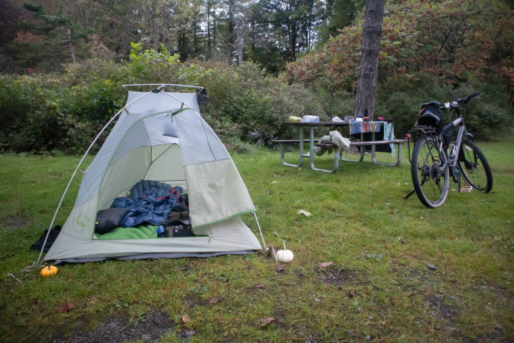 A tent sits in a grassy field. A bicycle is parked next to it, and bicycle bags can be seen inside the open tent flap. Camp cookware is on a picnic table behind the bicycle.