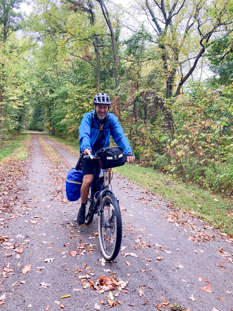 A man on a loaded touring bicycle rides towards the camera, on a wide trail through a forest with leaves on the ground. It may be lightly raining. The man is smiling.