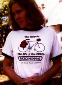 A woman, identified as Sandy Young, wears a t-shirt with a picture of a loaded touring bicycle. The shirt reads, "The Bicycle. The RV of the 1980s. BIKECENTENNIAL, The Bicycle Travel Association."