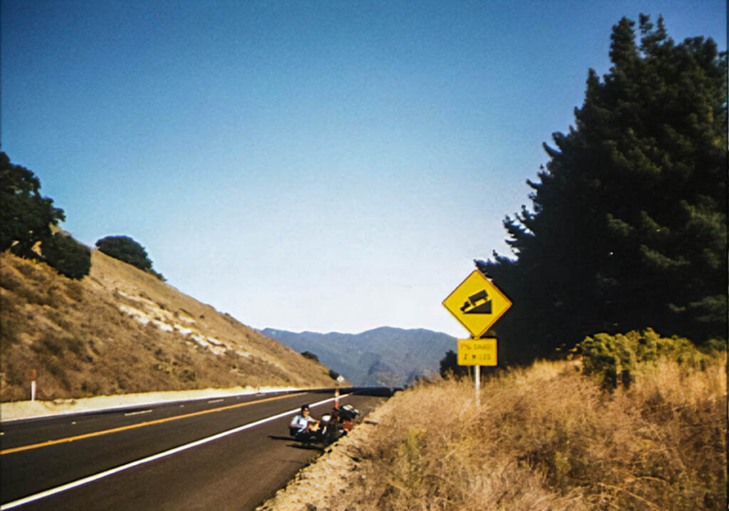 A bicyclist sits on the road at the top of a pass. A sign ahead of him warns of a 7% downgrade.