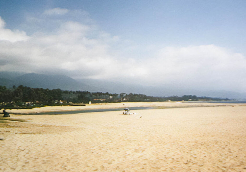 A wide, sandy beach sits under a blue sky with fluffy clouds. A vegetated hill rises from behind the beach.