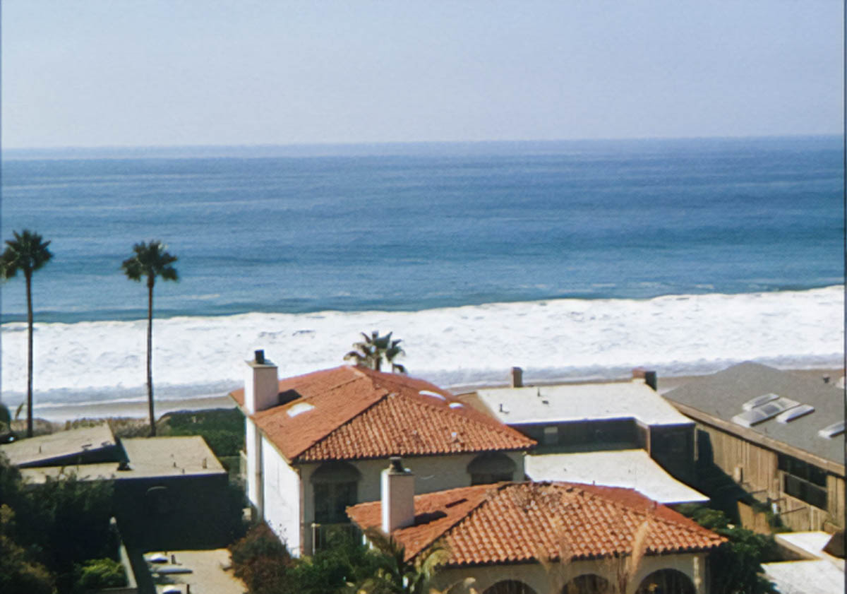 A blue ocean with waves breaking on a sandy beach. A large house is in the foreground, with tall palm trees off to the left.