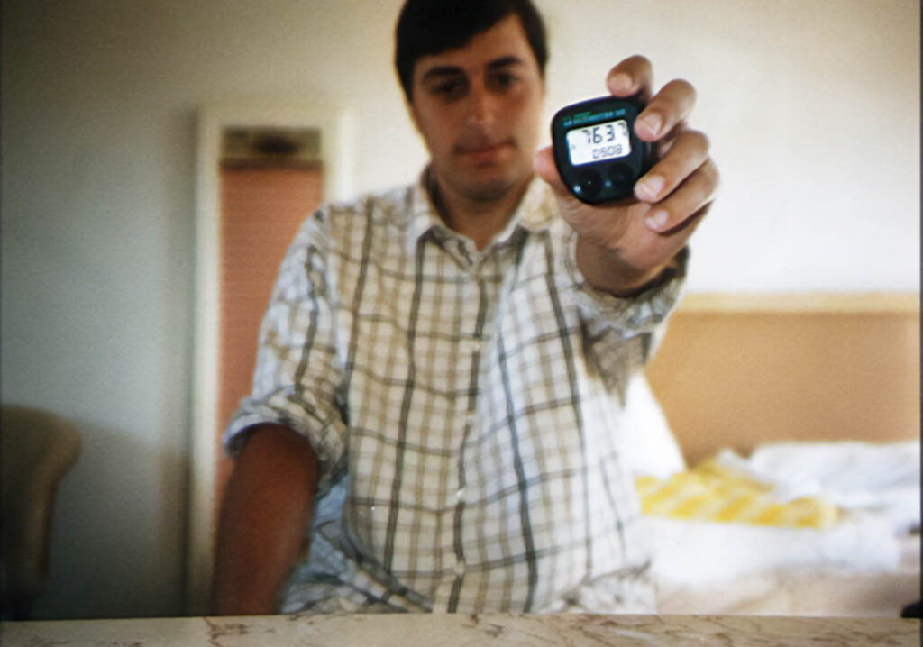 A man sitting in a motel room holds out a bike computer towards the camera. It reads "763.7".