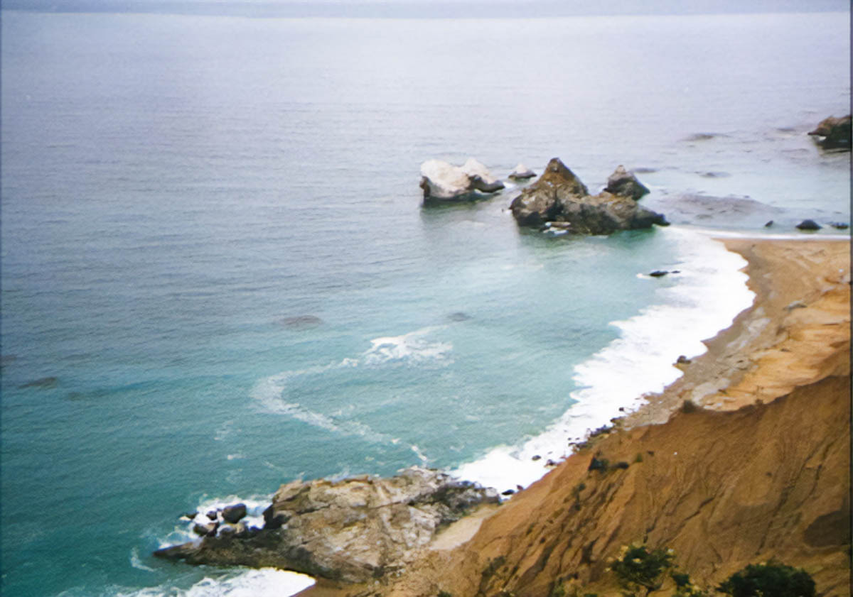 An outcropping splits a cove in two, with curved beaches on both sides of the outcropping. The water is turquoise blue.