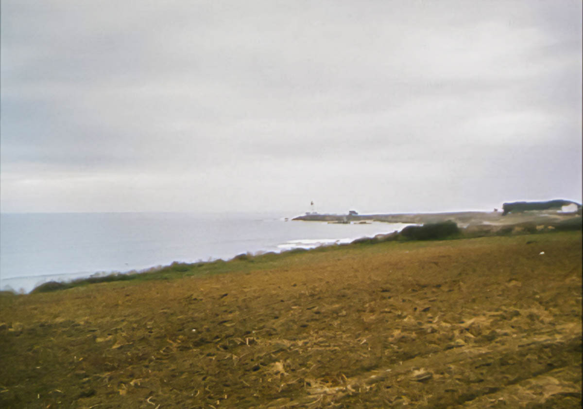 A harvested field in the foreground gives way to an ocean under a cloudy sky. In the distance, a lighthouse can be seen on the end of a point.