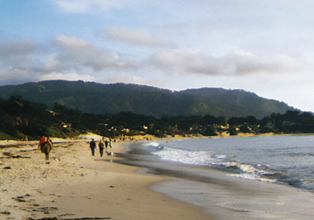 A curved shoreline forms a bay on a sandy beach. Many people stroll the beach, some with dogs. Sunset is nearing; fluffy clouds lit up by the sun float above a ridge of forested hills.