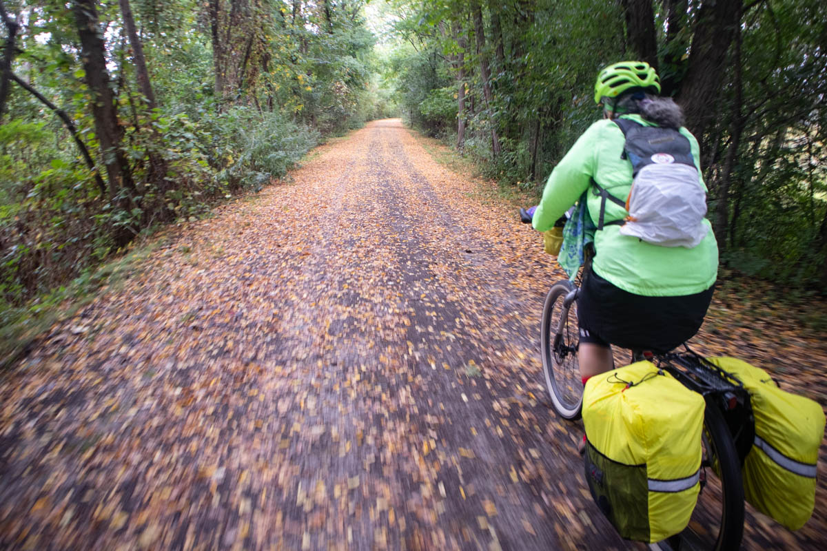 On a rainy day, a bicyclist with yellow pannier covers rides on a rail-trail through a forest. The pleasant-looking trail is mostly covered with brown and yellow leaves. The shot is probably taken from another bicycle; the pathway in the foreground is blurred by motion.