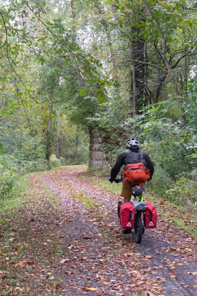 A bicyclist on a folding bike with full panniers, covered with red rain covers, rides on a leaf-covered rail trail through a forest.