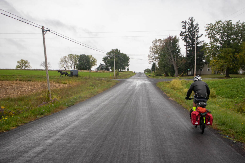 On a drizzly day, a bicyclist in rain gear with panniers rides through a rural landscape. Ahead of him is an Amish buggy that has just passed an intersection on a cross-street.