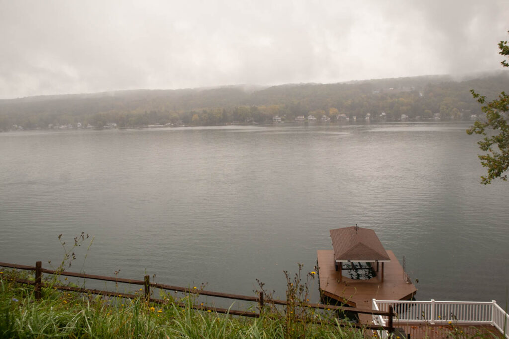 Clouds hang over a lake on a misty, rainy day. A fancy, covered private boat dock juts out into the lake in the foreground. The far side is wooded with autumnal trees.