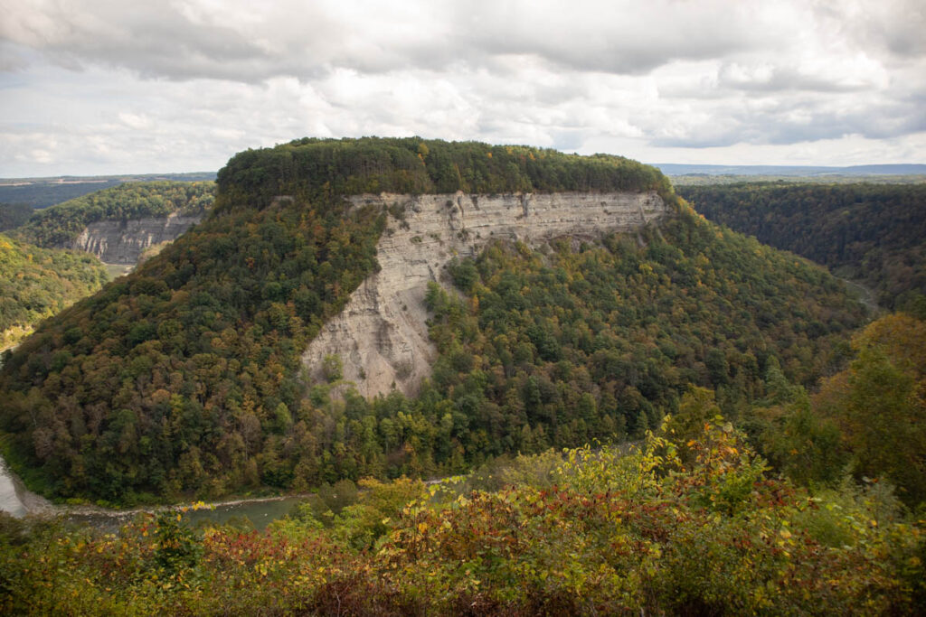 A river bends around an impressive granite mesa. The mesa and the banks of the river are lined with autumnal trees. The sky is mostly cloudy but the sun is peeking through in places.