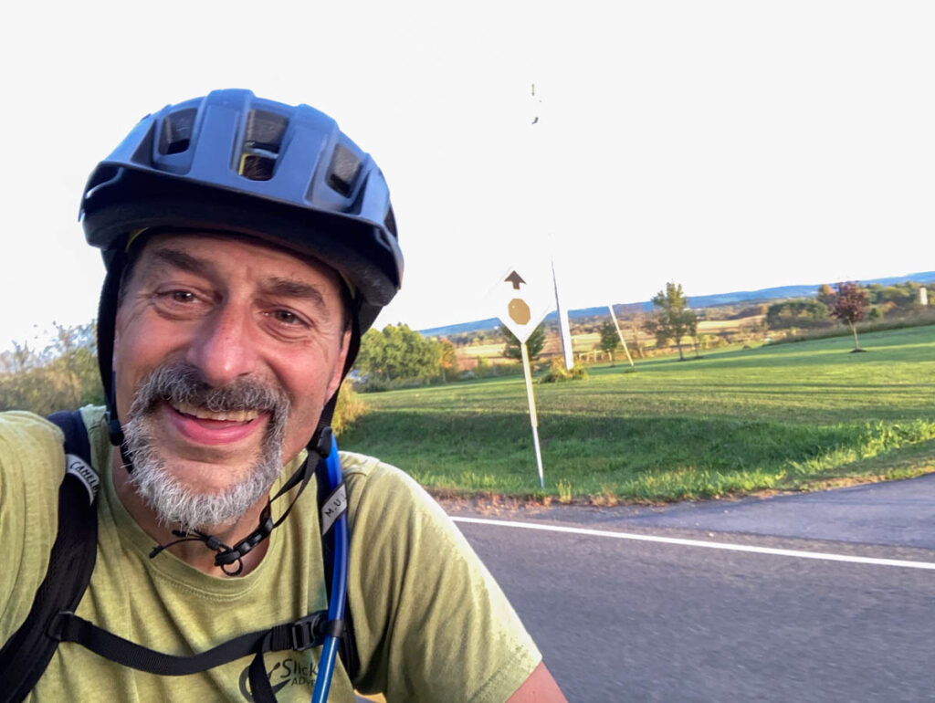 A smiling man, presumably riding a bicycle, takes a selfie with a late-afternoon rural landscape behind him