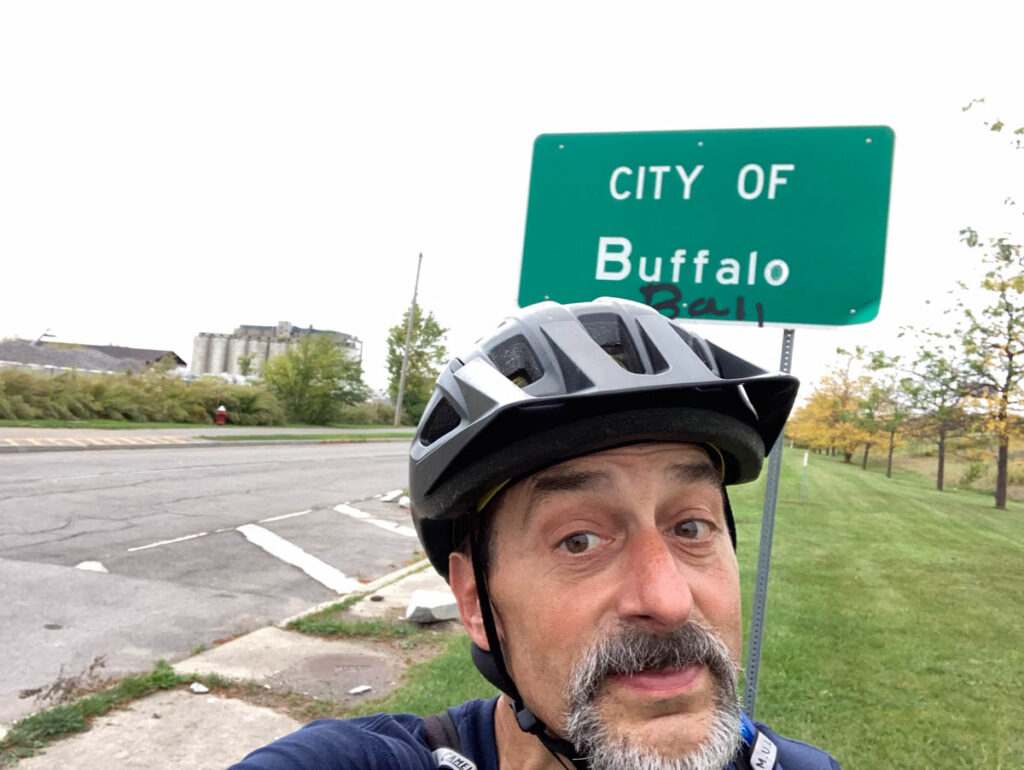 A man in a bicycle helmet stands in front of a green sign reading "CITY OF Buffalo". An abandoned factory can be seen in the background.