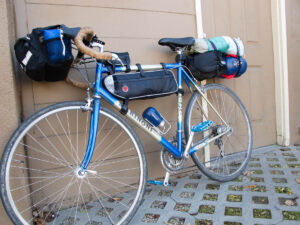 A somewhat vintage steel bicycle, painted blue and platinum and labeled as MARINONI, is loaded with a handlebar bag, a frame bag across the top tube, a small bag above the top tube, and a tent and sleeping bag strapped to a rear seatpost rack.
