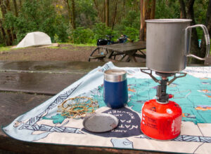 A campsite in a eucalyptus forest. It looks to have been recently raining. On the left a tent is set up, and a bicycle leans against a picnic table. In the foreground is a small camp stove with a pot above, along with a brass expanding coffee filter holder, and a thermal mug. They sit on top of a towel with a map of U.S. parks.