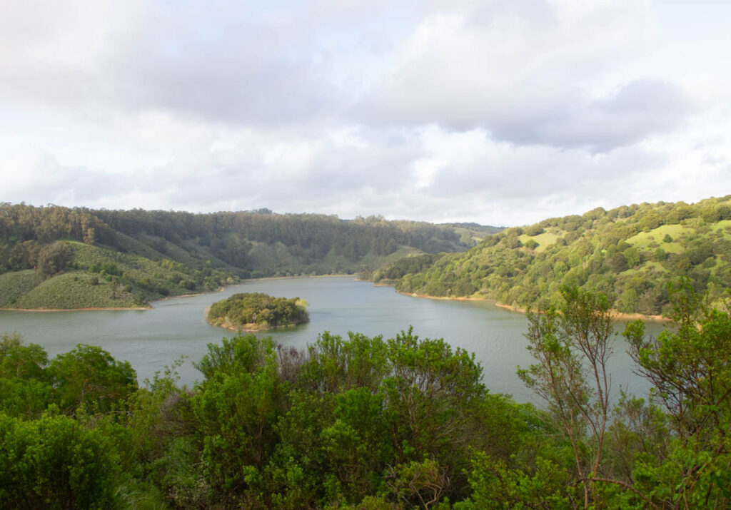 A mostly cloudy sky is seen over a lake nestled in green hills. Some of the hills are lit by a setting sun. A tree-covered island sits in the middle of the lake.