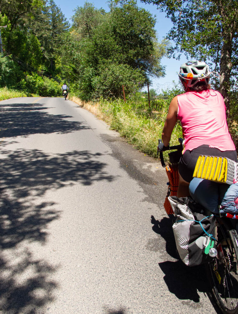 Two touring cyclists ride up a h ill on on a rural road. One is near the top of the hill, almost out of frame; the other is just in front of the camera, wearing a bright pink top, carrying full panniers and a sleeping pad.