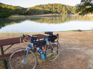 A bicycle loaded with bikepacking gear leans against a picnic table at a lakeside campsite. It appears to be early in the morning, with light falling on the trees and hills on the far side of the lake, reflected in the still lake waters.