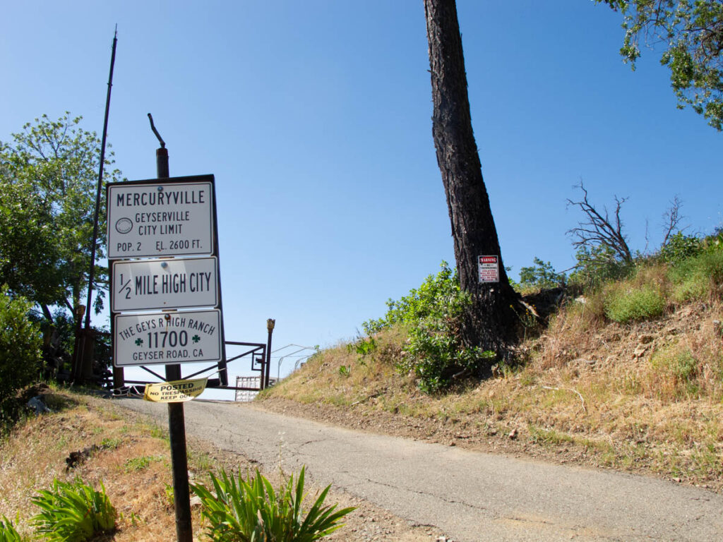 A dirt road in a dry landscape. A sign in the foreground reads "Mercuryville, half-mile high city. Elevation 2600 feet. Population 2. A sign nailed to a tree on the other side of the road reads "Warning, no trespassing. Violators will be shot. Survivors will be shot again!"