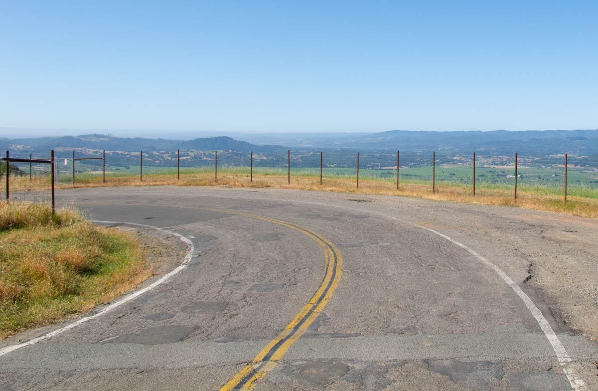 A road bends to the left high above a green valley. The sky is clear and blue. The road surface is somewhat broken up. A wire fence lines the road along the curve.