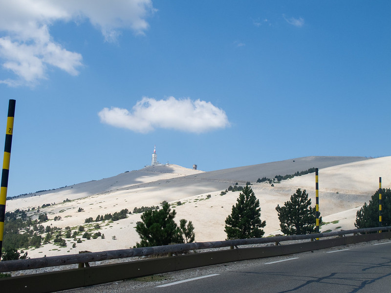 In the foreground is a road leading up to a barren mountaintop in the background. A snowplow pole in black and yellow, three meters tall is seen at the road's guardrail. The top of the mountain looks impossibly white but not snowy. A few clouds dot the sky above.