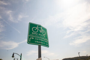 Against a sunny blue sky with wispy clouds, a green Bike Route sign reads "Three Rivers Heritage Trail: Great Allegheny Passage to Washington, D.C. 350m"