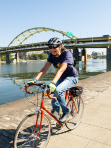 A smiling woman rides an orange bike along a waterfront pathway, with two yellow steel bridge in the background.