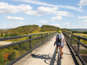A woman on a bicycle rides across a high bridge over a highway. The landscape is rolling hills with trees, some of which are yellow and red. The sky is blue with fluffy clouds. On the ridge ahead is a wind farm.