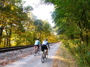 Two cyclists ride away from the camera side-by-side in yellow morning light on a gravel path along a rail line. A few leaves are on the ground. The path extends straight off into the distance.
