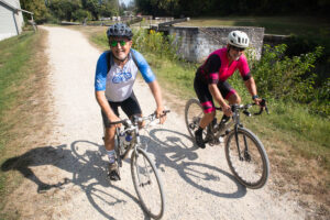 Two bicyclists ride towards the camera on a gravel path. One, wearing a Climate Ride jersey, looks at the gamera and smiles.