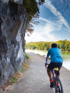 A bicyclist rides away from the camera along a gravel trail. On the left side of the trail is a sheer rock face; on the right side is a river. The other side of the river has a forest with fall colors.