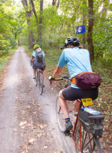 Two women on bicycles ride away from the camera on a gravel pathway through a green forest. Some brown leaves are on the ground. The women's helmets both have small flags attached, with an image of the earth from space.