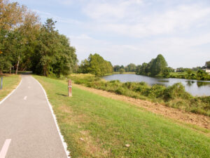 A two-way bike path meanders through a grassy area, entering a wooded area up ahead. To the right is seen a small river.