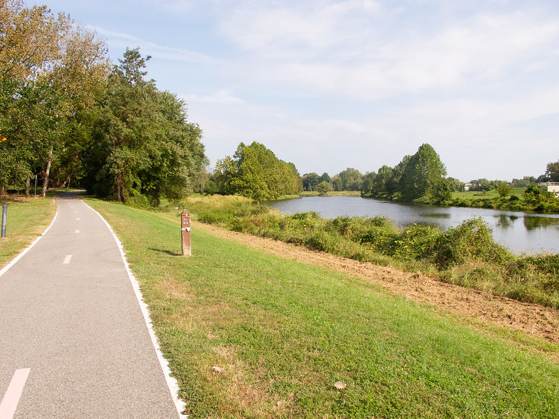 A two-way bike path meanders through a grassy area, entering a wooded area up ahead. To the right is seen a small river.