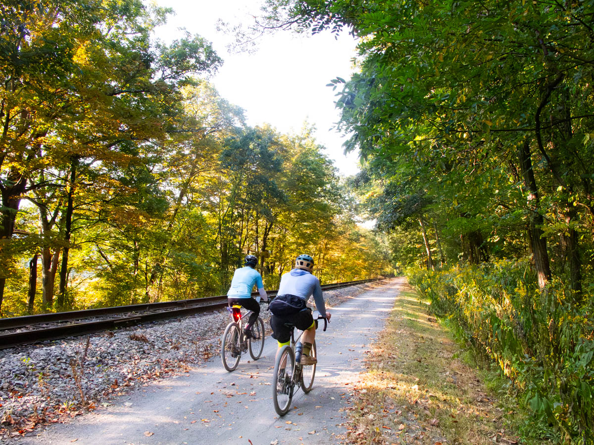Two cyclists ride away from the camera side-by-side in yellow morning light on a gravel path along a rail line. A few leaves are on the ground. The path extends straight off into the distance.