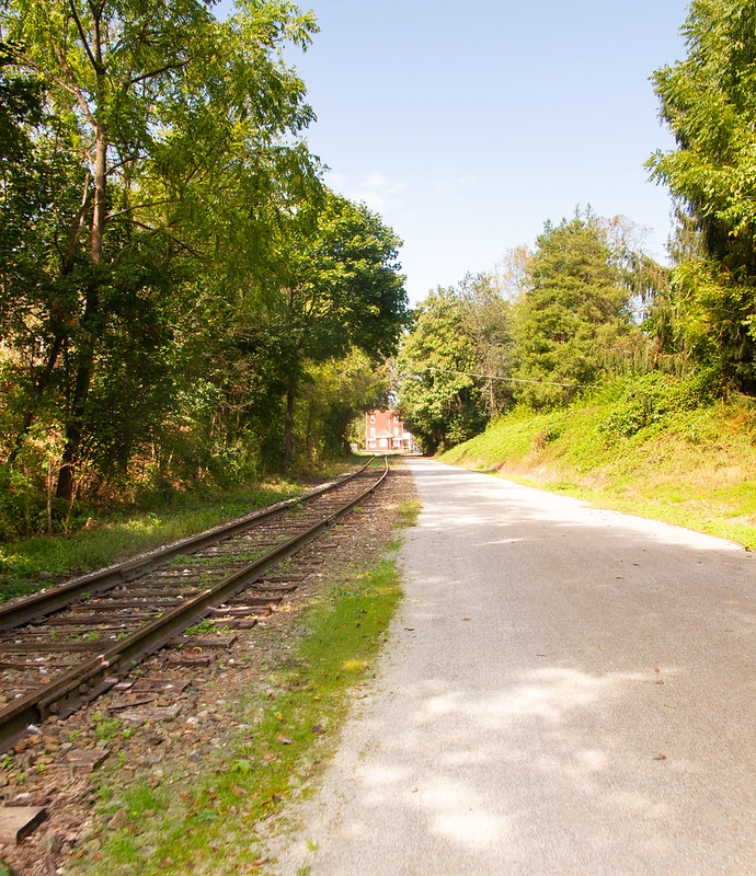 A gravel pathway parallels a railroad line. The railroad has some plants growing between the ties, suggesting it is lightly used. In the distance, the pathway goes under an arch of trees; a red building is seen past the trees.