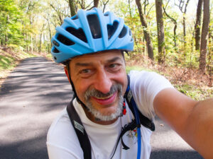 A smiling man in a bicycle helmet holds out the camera ahead of him for a selfie. The road behind curves through a bright forest, quiet and dotted with fallen leaves.